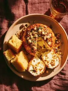 A golden-baked Aruban keshi yena cheese dome topped with nuts and herbs, served with cornbread slices on a tan ceramic plate, under soft afternoon light.