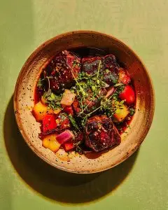 A rustic bowl of Barbadian lamb chops in tomato and onion sauce with thyme, bell peppers, and herbs, placed on a muted green surface under warm daylight.
