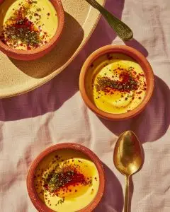 Terracotta bowls of Bengali mishti doi, golden sweet yogurt topped with crushed nuts and saffron, on a soft pink cloth beside brass spoons in warm daylight.