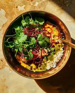 A brass bowl filled with Bangladeshi bhuna dal khichuri — golden lentils, rice, and spices — garnished with coriander and herbs, photographed in warm sunlight.