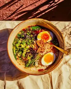 A ceramic bowl of Filipino arroz caldo chicken rice porridge topped with soft-boiled eggs, chili, and spring onions, photographed in warm daylight on a pale cloth background.