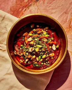 A close-up of a brass bowl filled with rich orange-red Indian carrot halwa topped with sliced pistachios, almonds, and carrot slivers, photographed on a peach-pink cloth under warm natural light.