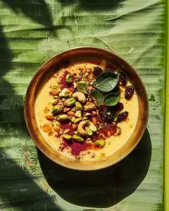 A bowl of creamy South Indian paal payasam rice pudding with saffron and chopped pistachios, almonds, and raisins, placed on a green banana leaf in daylight.
