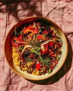 A ceramic bowl of Nigerian jollof rice made with red bell peppers, onions, and tomatoes, garnished with herbs, set on a soft pink cloth in bright daylight.