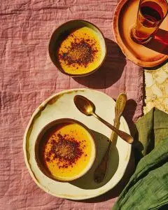 Two small ceramic bowls of golden-brown Caribbean sweet potato pudding sprinkled with cinnamon, styled on a rose-pink linen cloth in soft sunlight.